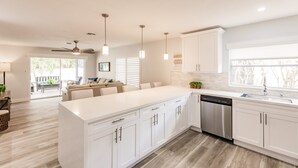 A modern kitchen with white cabinetry, a stainless steel dishwasher, pendant lighting, and a large window providing natural light. - Serenity by the Sea: Heated Pool, Bikes & Coastal Charm (Naples)