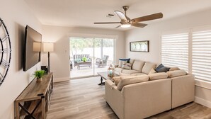 A modern living room with a sofa, a TV, and a ceiling fan. - Serenity by the Sea: Heated Pool, Bikes & Coastal Charm (Naples)