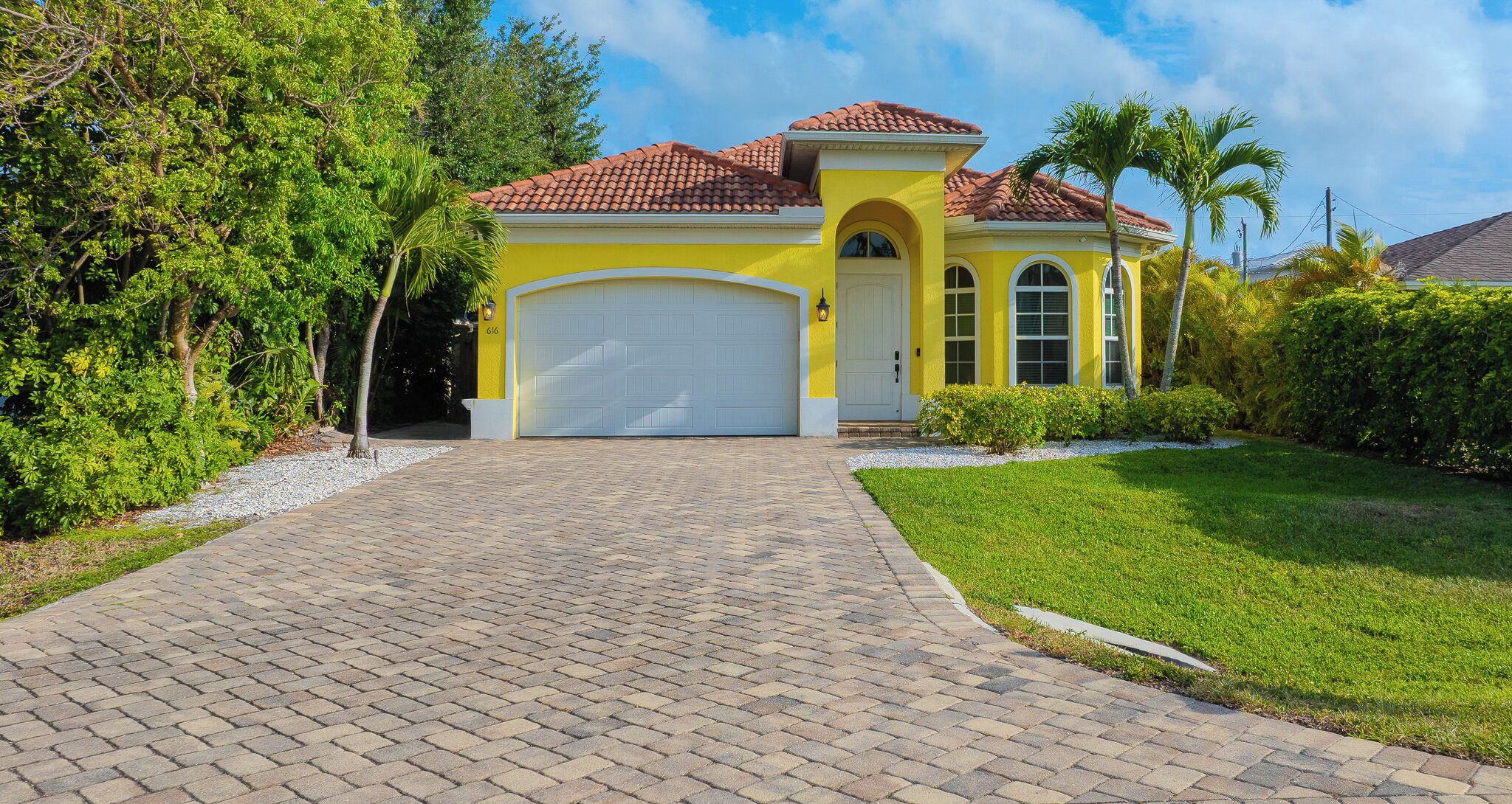 A yellow house with a red-tiled roof, a white garage door, and a paved driveway leading to the entrance.