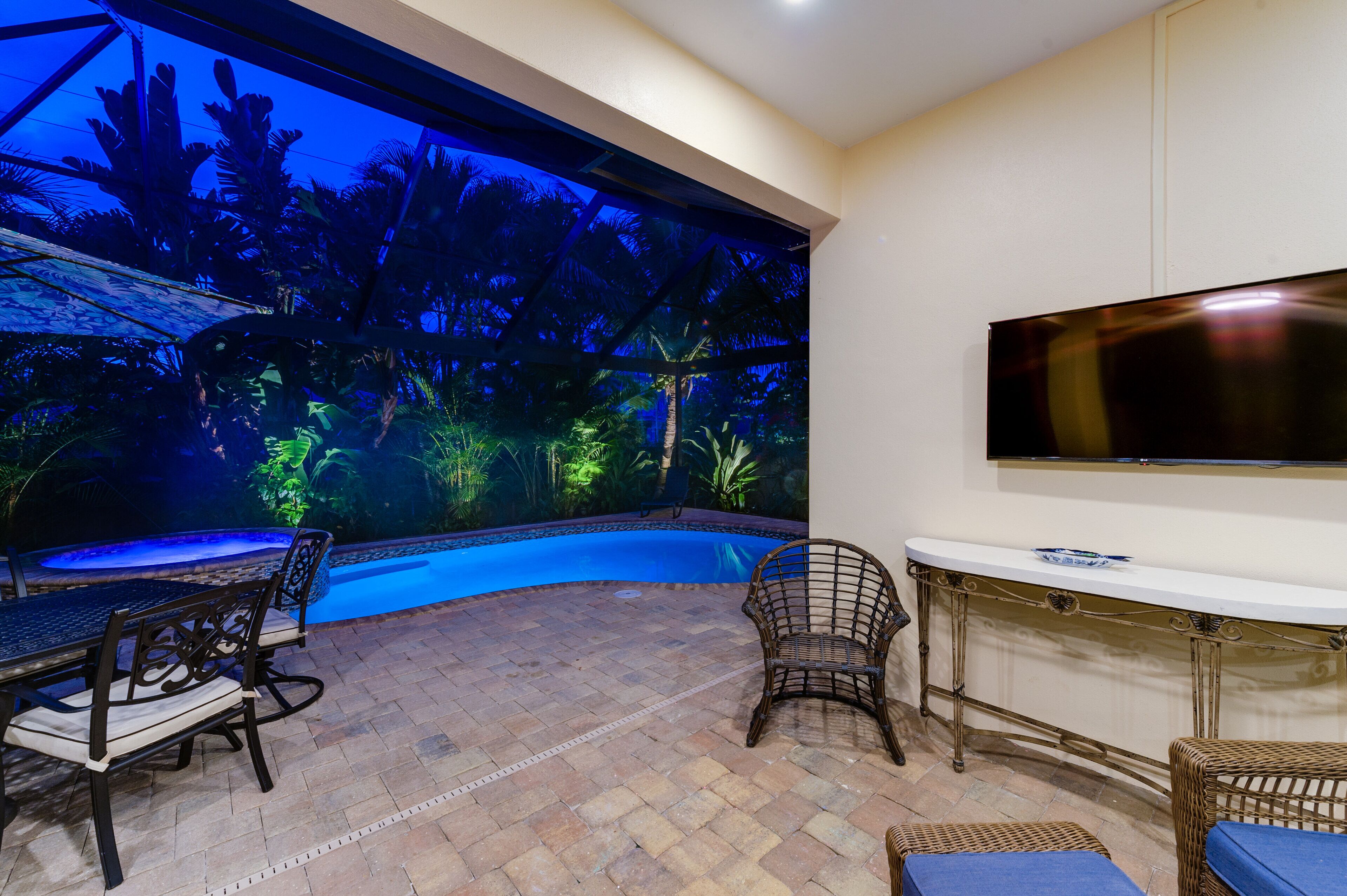 Una zona de piscina amb una estructura de vidre, un televisor fixat a la paret i mobiliari exterior.