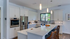 Une cuisine moderne avec des meubles blancs, des appareils en acier inoxydable, un grand îlot central doté de tabourets et un éclairage suspendu.