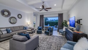 A modern living room with a grey sofa, armchairs, a coffee table, and a TV. There are decorative mirrors on the wall and a ceiling fan. - Casa Allegra | Bright, Modern 4 BR Pool Home in Naples Park (Naples)