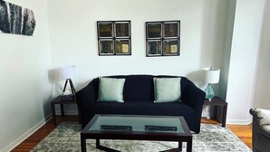 Ein Wohnzimmer mit einem dunklen Sofa, zwei Beistelltischen, einem Glastisch und einem gemusterten Teppich.