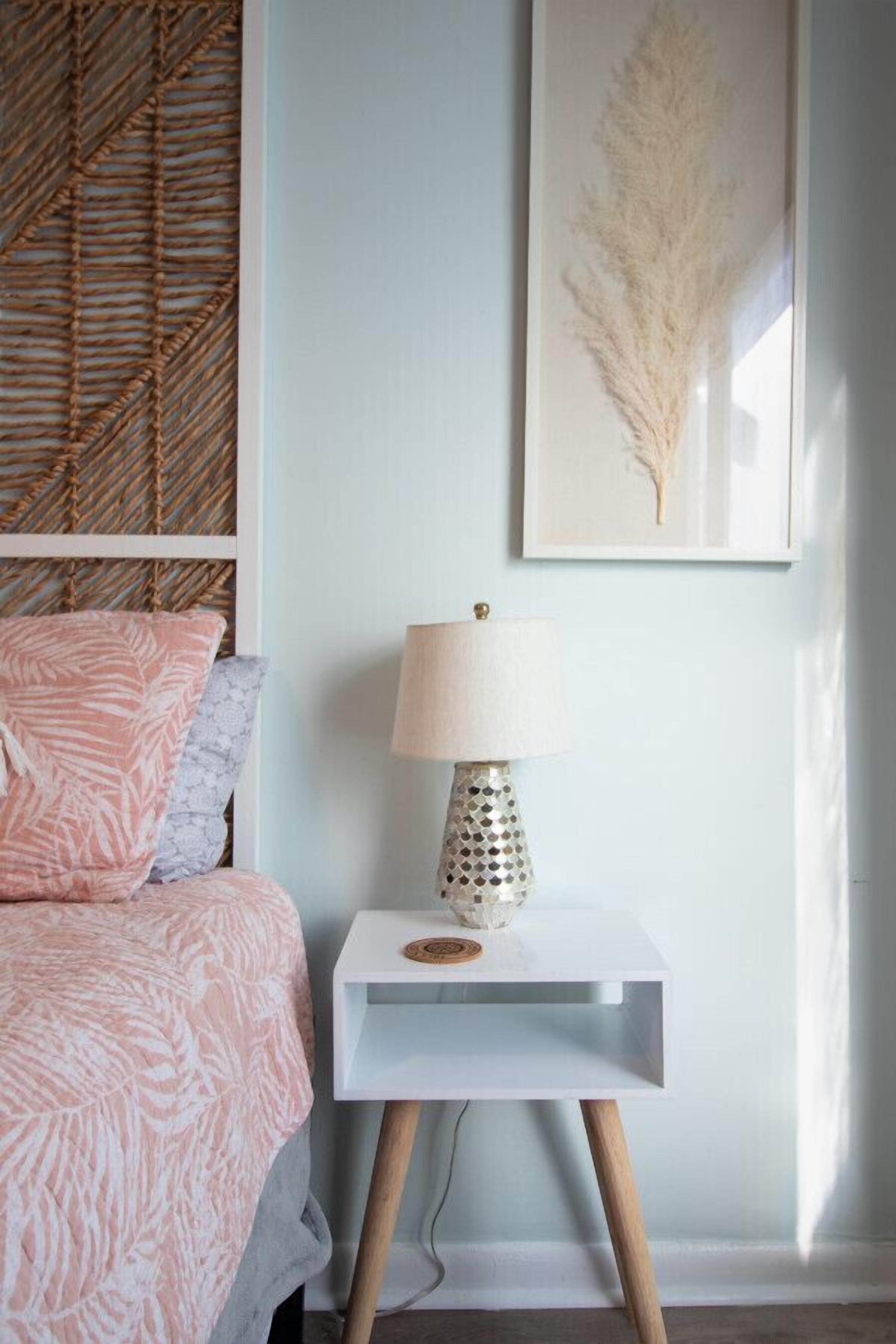 Une chambre à coucher avec un lit, une table de chevet avec une lampe, une œuvre d’art encadrée au mur et une tête de lit en osier.