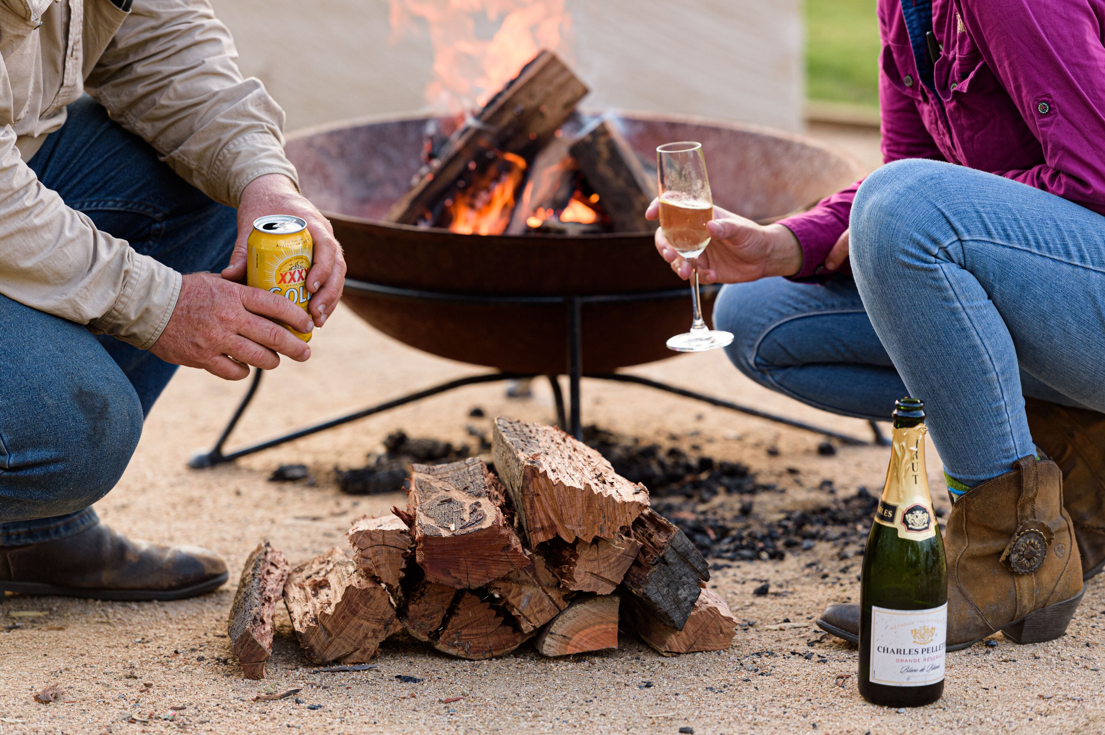 A couple sitting by a fire pit with a bottle of champagne and a can of beer.