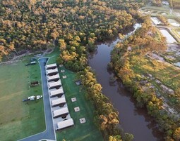 Vista aérea de un camping con vehículos recreativos estacionados a lo largo de una carretera, rodeado de un bosque denso y un río.