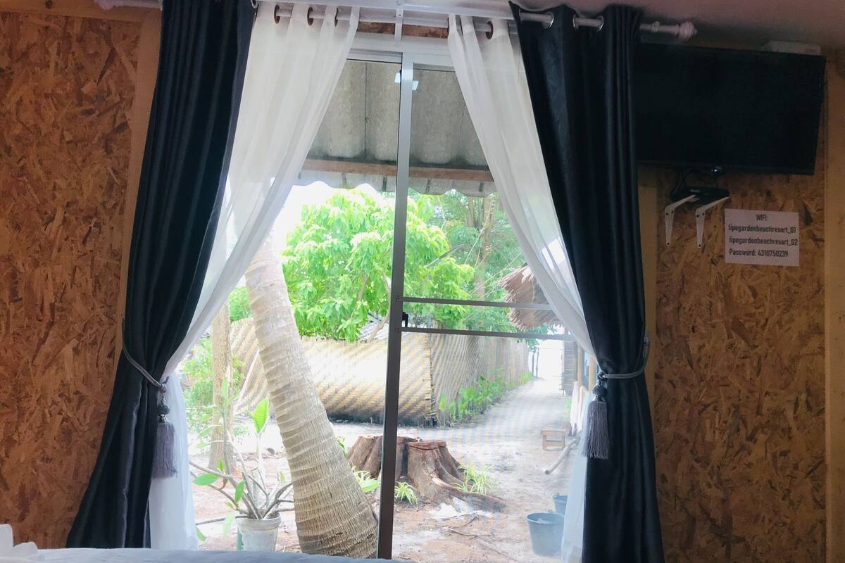A room with a bed, a window with white curtains, a sign with WiFi details, and a view of a garden.