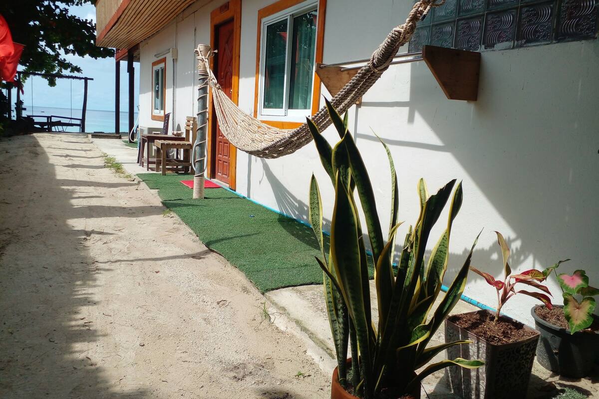 A building with a sandy pathway leading to a door, a hammock hanging from a post, and potted plants along the wall.