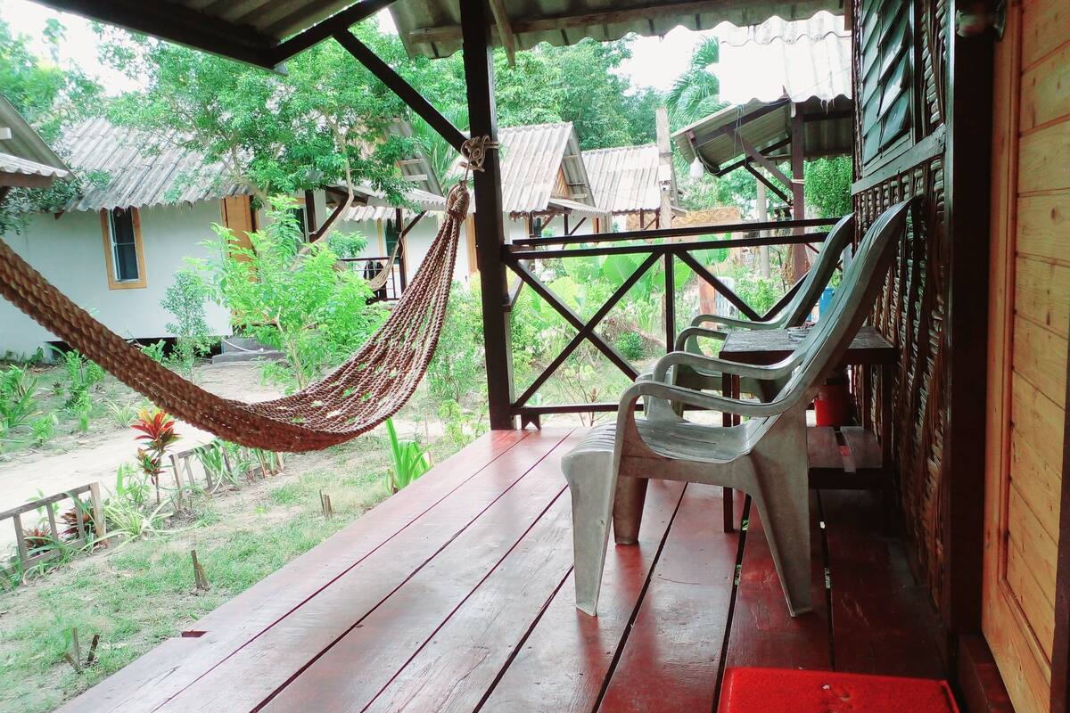 A wooden deck with a hammock and plastic chairs, overlooking a garden with various plants and a building with a corrugated metal roof.