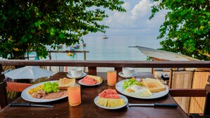 Una mesa de madera con platos de desayuno, un vaso de jugo y vista al mar con barcos.