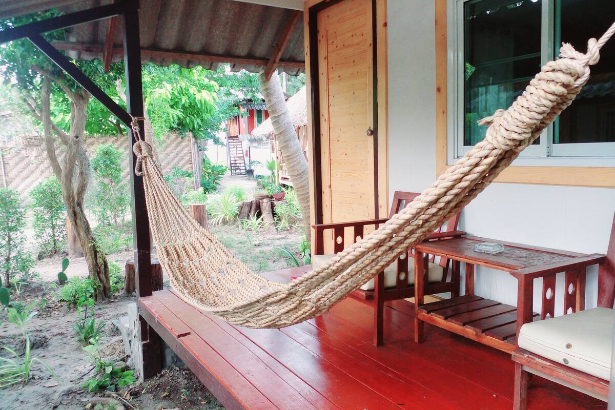 A hammock on a wooden deck with a view of a garden and a building in the background.