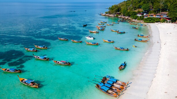 Una playa con numerosas embarcaciones ancladas cerca de la orilla, rodeada de exuberante vegetación y un mar azul cristalino.