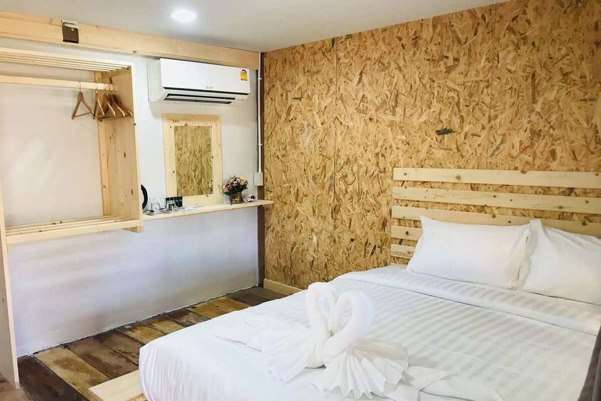 A hotel room with a wooden headboard, a bed with white bedding, a wooden floor, a wooden wall with a textured pattern, a mirror, and an air conditioning unit.