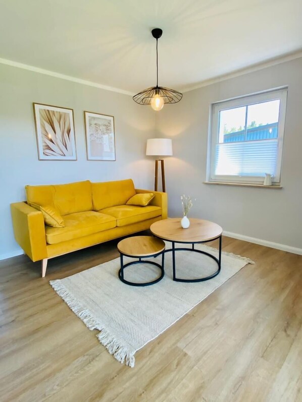 Quarto com sofá amarelo, mesa de madeira redonda, tapete branco e uma luminária.