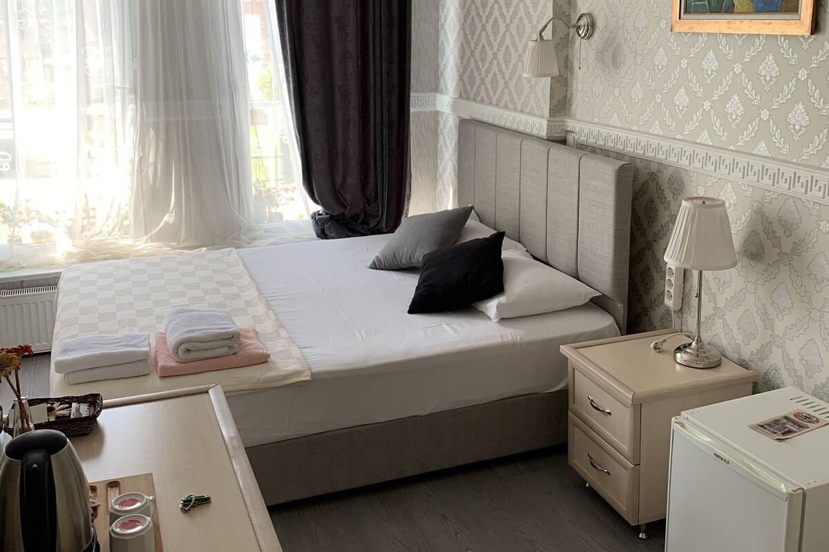 Habitación de hotel con una cama grande, una mesita de noche con lámpara, un refrigerador pequeño y una ventana con cortinas traslúcidas.