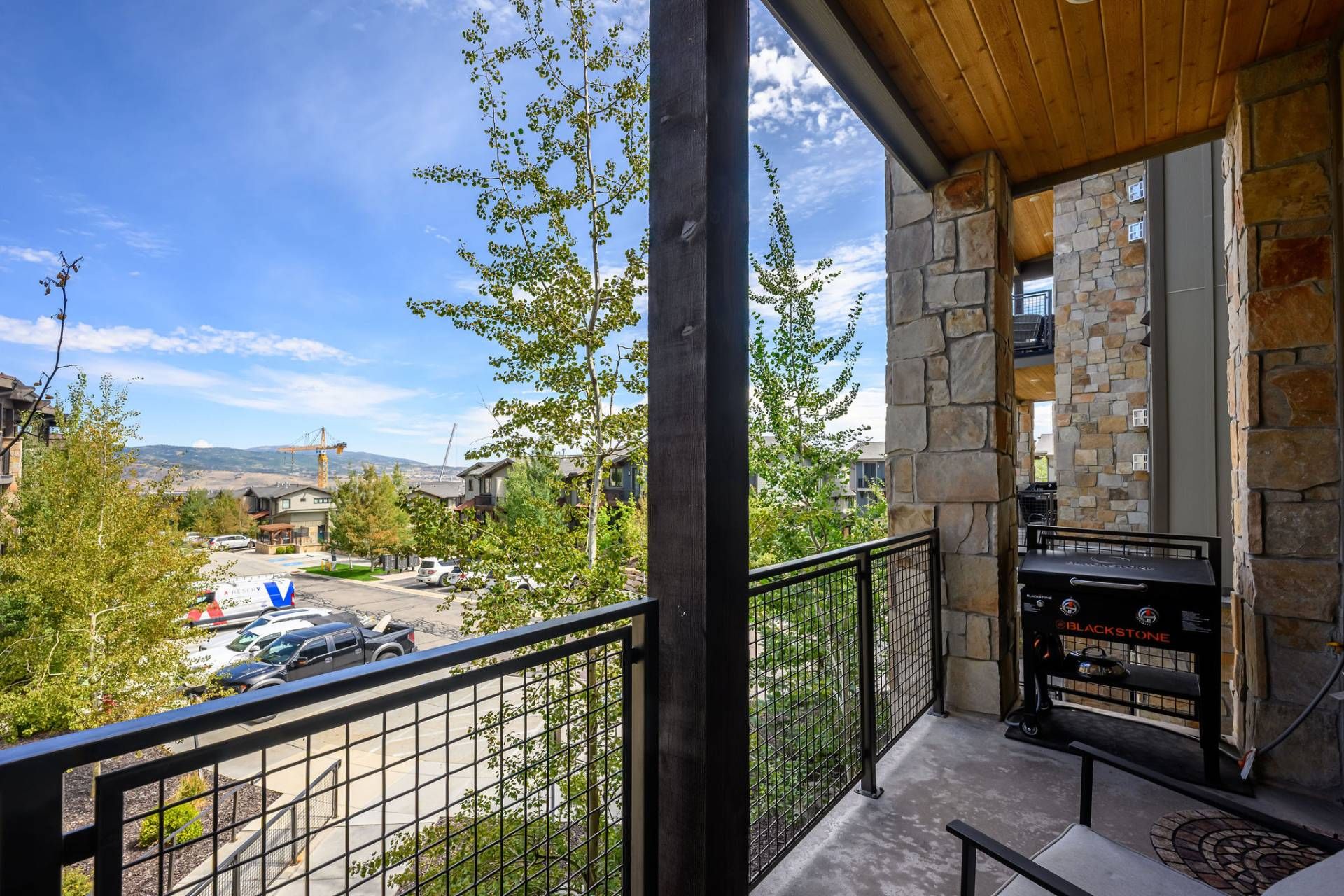 Un balcón con muro de piedra, una parrilla y vistas a una zona residencial con coches y árboles.