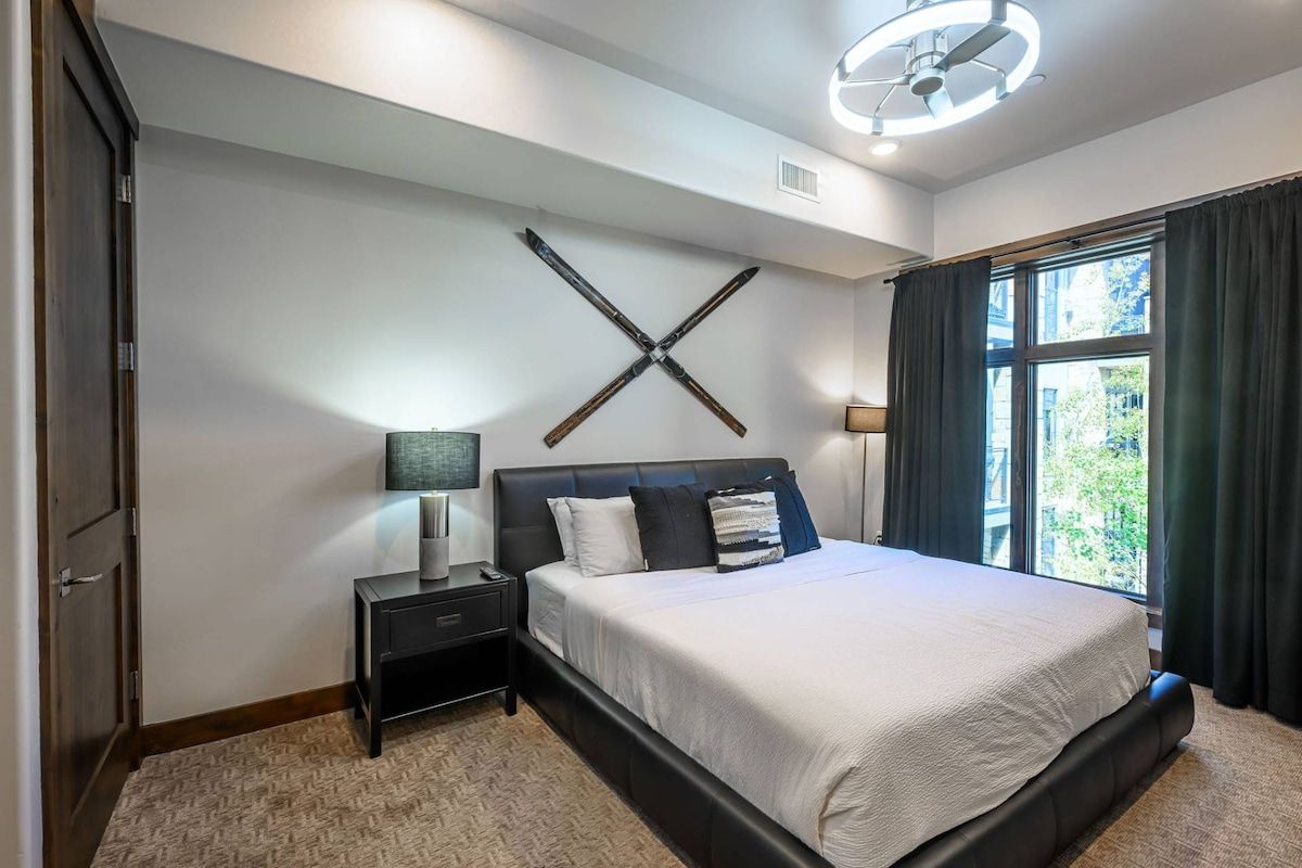 A modern bedroom with a large bed, a nightstand, a wall-mounted light fixture, and a window with curtains.