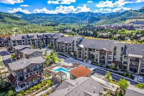 A residential area with modern multi-story buildings, a central swimming pool, and a mountainous backdrop.