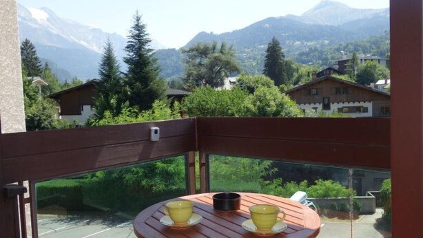 Een balkon met een tafel en stoelen, met uitzicht op een bergachtig landschap met huizen en begroeiing.