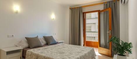 Un dormitorio con cama, mesitas de noche, una planta en maceta y un balcón con cortinas.