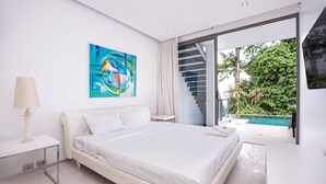 Een moderne slaapkamer met een groot bed, een bureau, een lamp, een schilderij en uitzicht op een zwembad en groen.