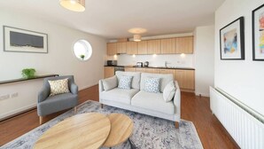 Un apartamento moderno con un sofá gris, una mesa de centro redonda, una cocina con armarios de madera y un ventanal pequeño y circular.