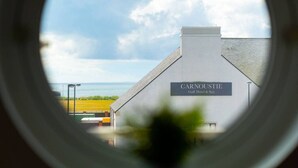 Vista a través de una ventana circular de un edificio con la leyenda “CARNOUSTIE Golf Hotel & Spa”, con un cielo despejado y un cuerpo de agua al fondo.