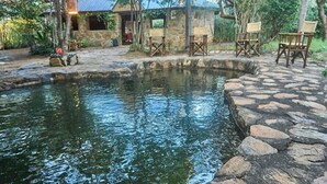 A stone patio with a small pond, wooden chairs, and a rustic stone building with a thatched roof. - Tindiga Tented Camp (Mangola)