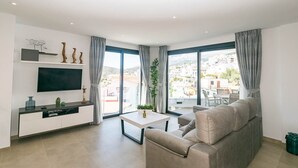 Sala de estar moderna com sofá, TV e varanda com vista para prédios e colinas.