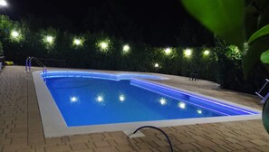 Een zwembad met blauwe LED-verlichting langs de randen, omgeven door een tegel terras en een haag.
