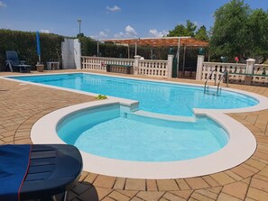 A rectangular swimming pool with a small hot tub, surrounded by a brick deck and a decorative fence.