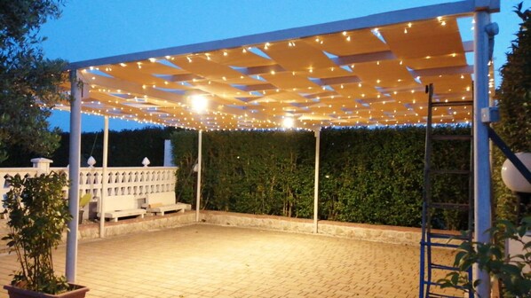 A covered patio area with string lights, a white fence, and a hedge in the background.