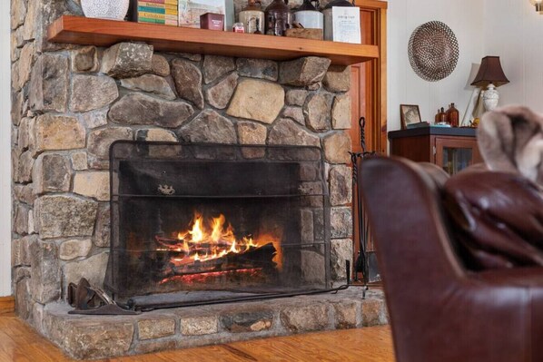 Una chimenea de piedra con fuego encendido, un estante superior con libros y objetos decorativos, y un sillón de cuero marrón en primer plano.