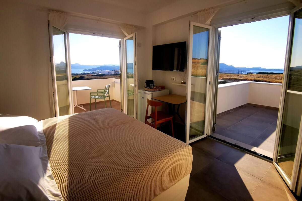 A hotel room with a bed, a desk, a chair, and a TV. There is a balcony with a view of the landscape.