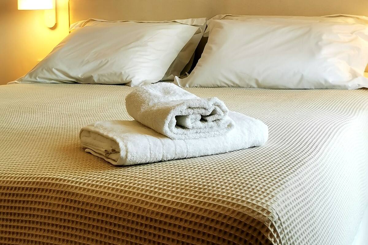 A neatly made bed with white linens and a folded towel, a wall-mounted light, and three framed shell decorations.