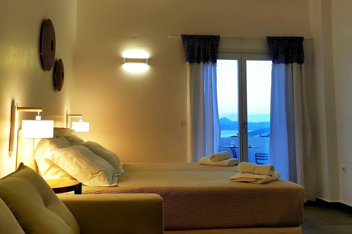 A bedroom with a bed, a sofa, a window with curtains, and a view of the sea.