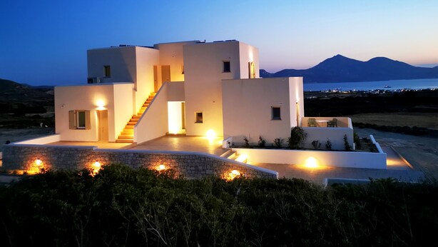 A modern, multi-story building with a staircase leading to the upper levels, surrounded by greenery and a view of the sea and mountains in the background. - Kallisto Rooms (Milos)