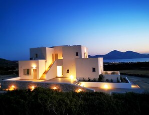 A modern, multi-story building with a staircase leading to the upper levels, surrounded by greenery and a view of the sea and mountains in the background. - Kallisto Rooms (Milos)