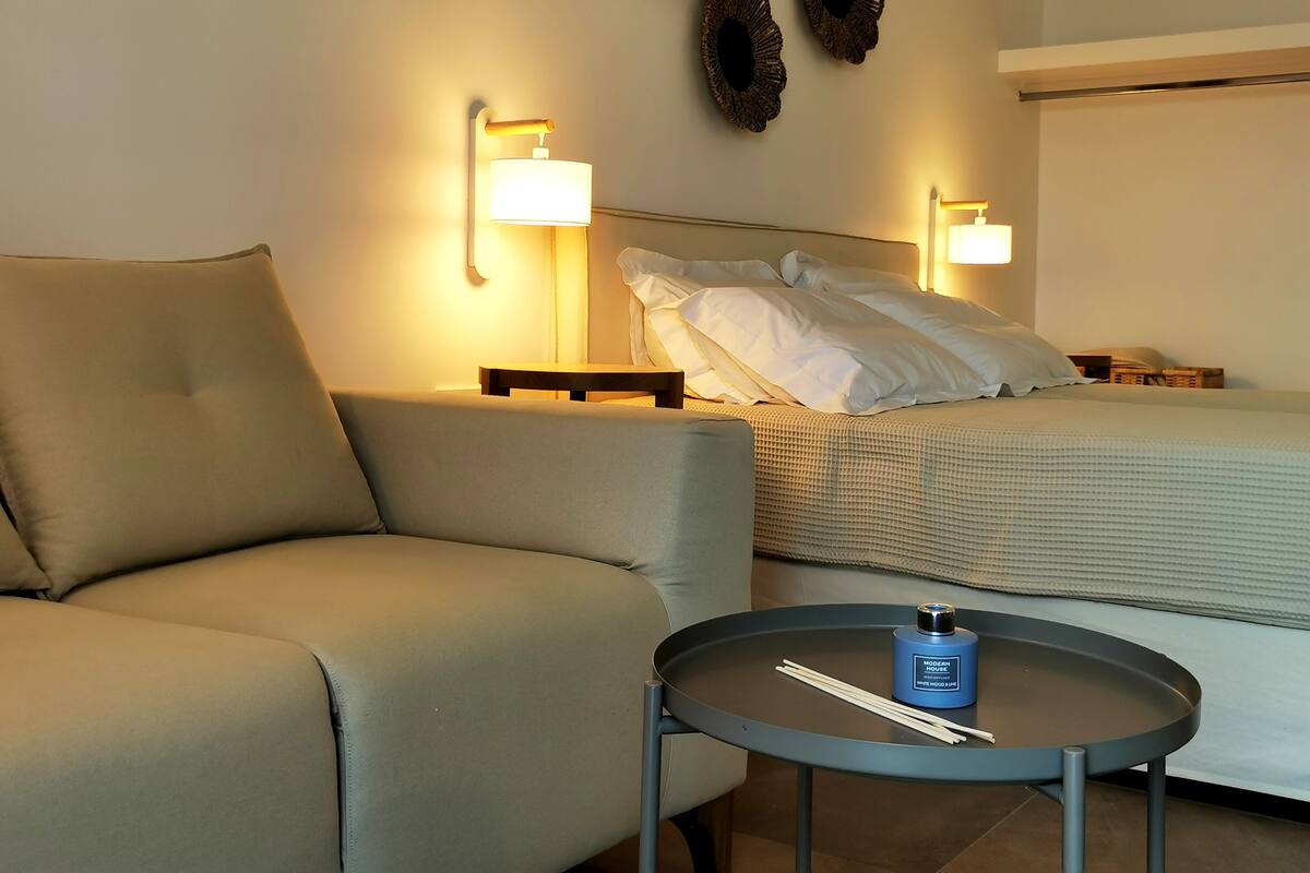 A modern hotel room with a sofa, a round table with a bottle of Modern House White Wood incense, and a bed with white bedding.