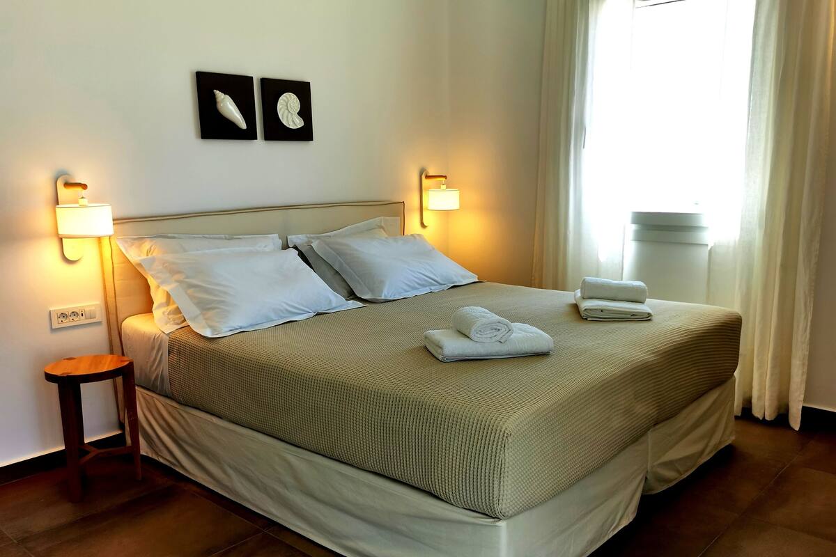 A neatly made bed with a beige bedspread, two pillows, and a rolled towel on the side. There are wall-mounted light fixtures on either side of the bed, and a small wooden stool is visible to the left.