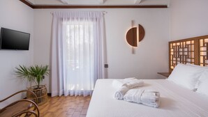 Habitación de hotel con una cama grande, un televisor de pantalla plana en la pared, una planta en maceta, un cabecero de madera y un ventanal con cortinas traslúcidas.