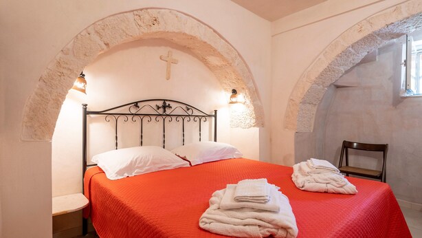 A bed with a red bedspread, white pillows, and a cross above the headboard. There are two folded towels on the bed.