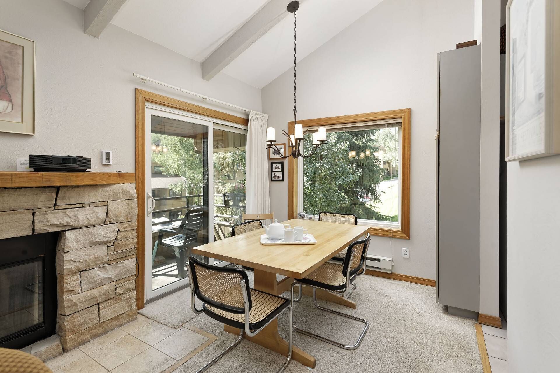 Un comedor con chimenea de piedra, una mesa de madera y sillas. Se ve una ventana con vistas a árboles y una lámpara colgante sobre la mesa.