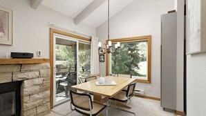 Un comedor con chimenea de piedra, una mesa de madera y sillas. Se ve una ventana con vistas a árboles y una lámpara colgante sobre la mesa.