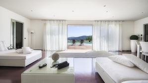 Un soggiorno moderno con un grande tavolino in vetro, due divani bianchi e vista su una piscina e montagne.