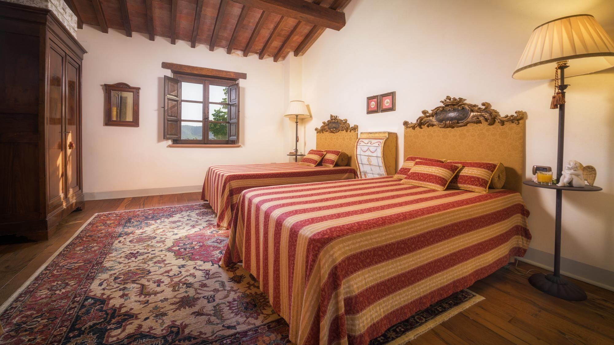A room with two beds, a wooden floor, a patterned rug, a window, and a lamp.
