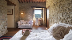 Una stanza accogliente con parete in pietra, un divano bianco, un letto con copripiumino fantasia e vista sul verde attraverso una finestra.