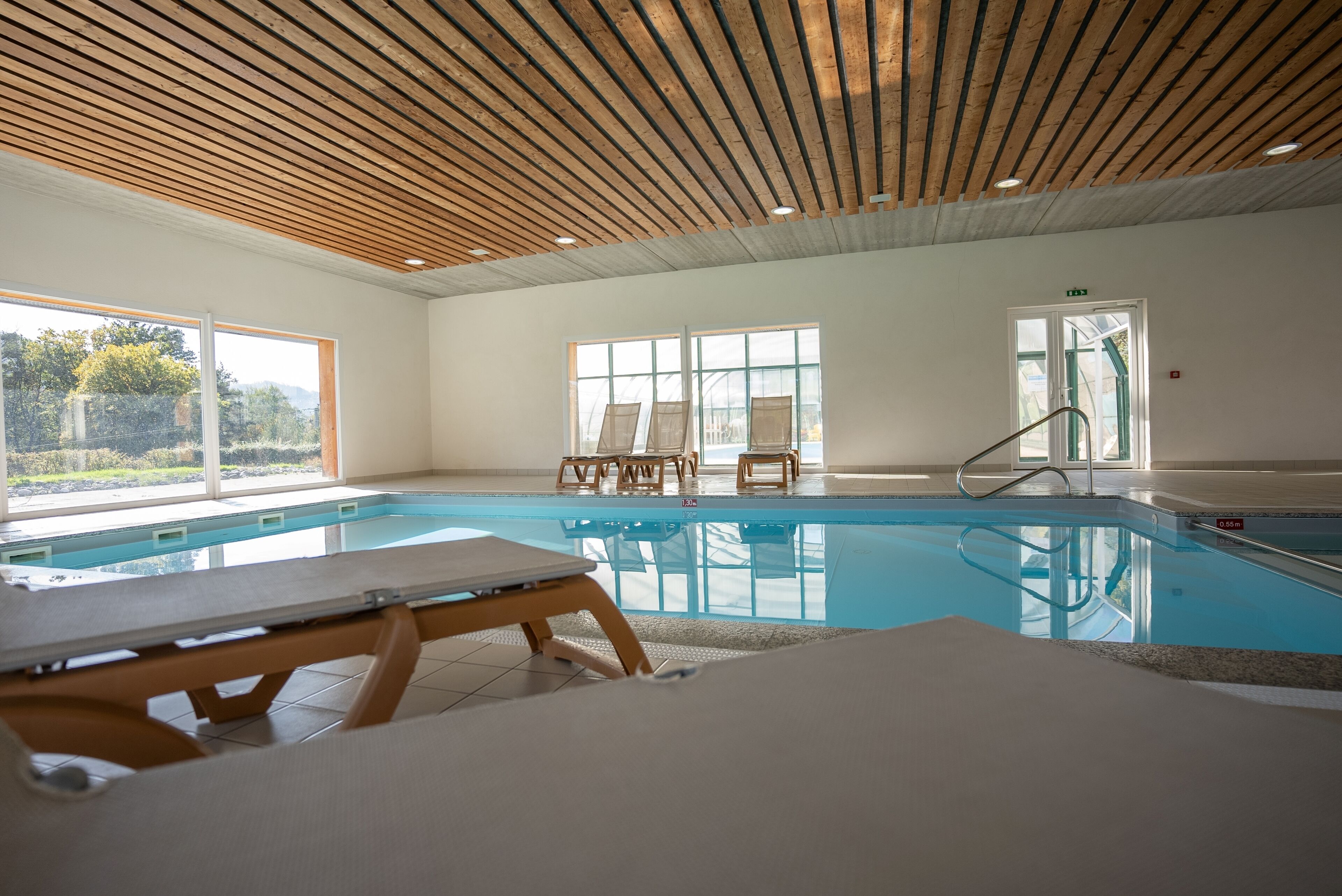 An indoor swimming pool with wooden ceiling beams, large windows, and lounge chairs.