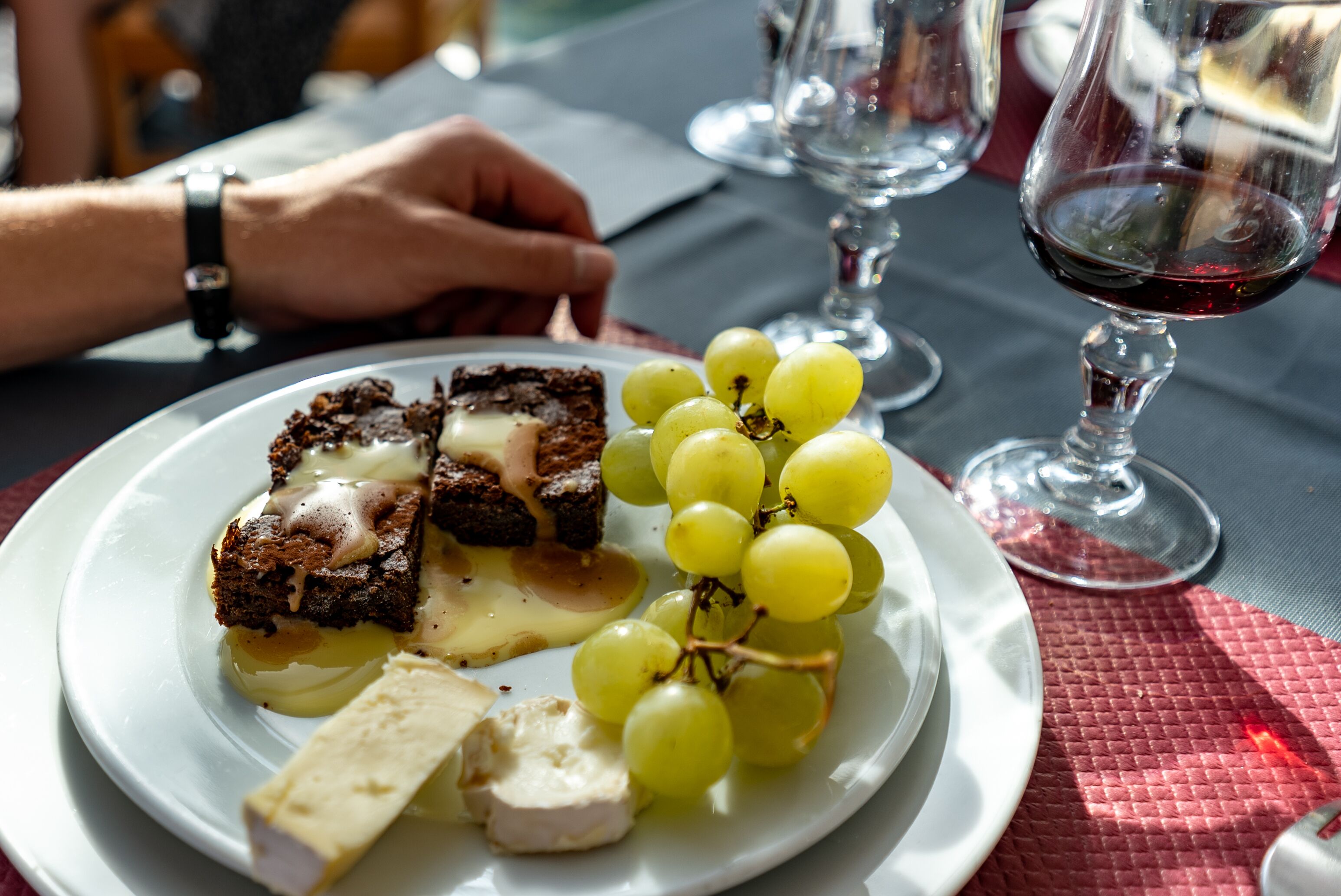 A plate with a dessert consisting of a chocolate cake with a creamy topping, a piece of white cheese, and a bunch of green grapes.