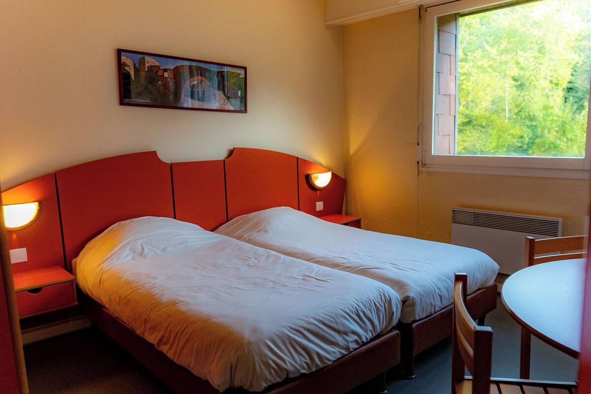 Cama doble con cabecera roja, dos mesitas de noche con lámparas, una mesita pequeña y ventana con vistas a zonas verdes.